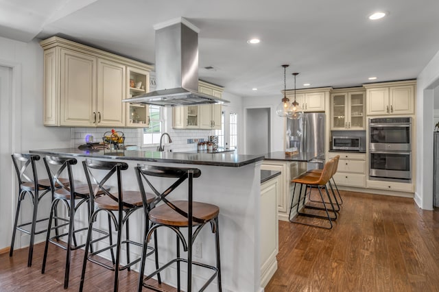 kitchen with island exhaust hood, cream cabinetry, a breakfast bar area, pendant lighting, and appliances with stainless steel finishes