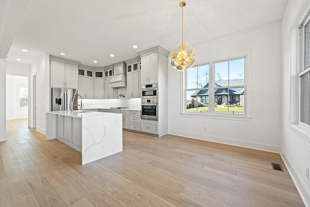 kitchen with light hardwood / wood-style flooring, an island with sink, decorative light fixtures, custom exhaust hood, and appliances with stainless steel finishes