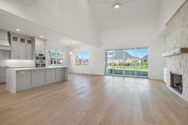 unfurnished living room with a stone fireplace, high vaulted ceiling, ceiling fan with notable chandelier, and light wood-type flooring