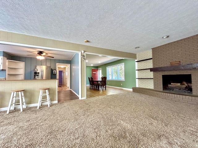 unfurnished living room featuring a brick fireplace, a textured ceiling, light colored carpet, and ceiling fan