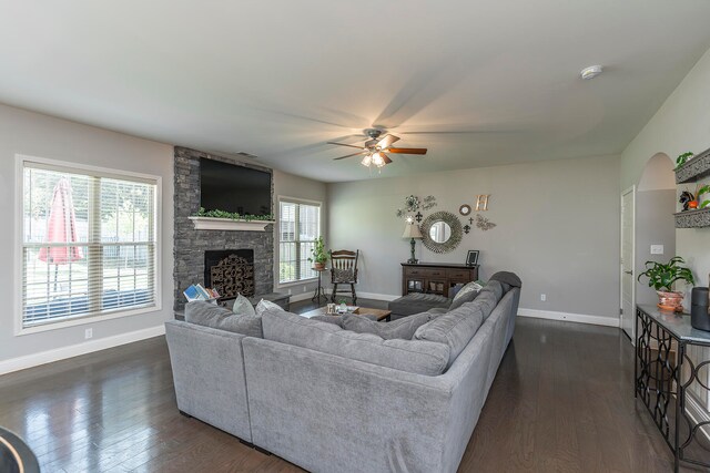 living room with dark wood-type flooring, ceiling fan, a healthy amount of sunlight, and a stone fireplace