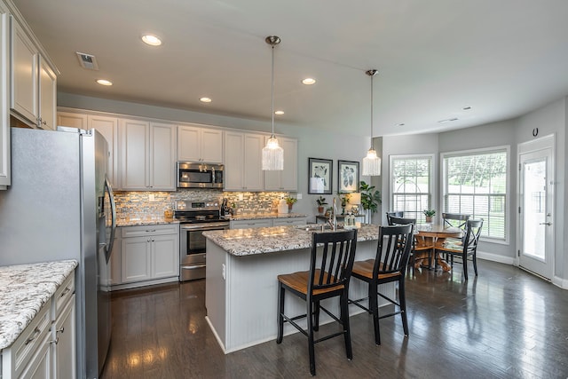 kitchen with dark hardwood / wood-style floors, stainless steel appliances, decorative light fixtures, and a center island with sink