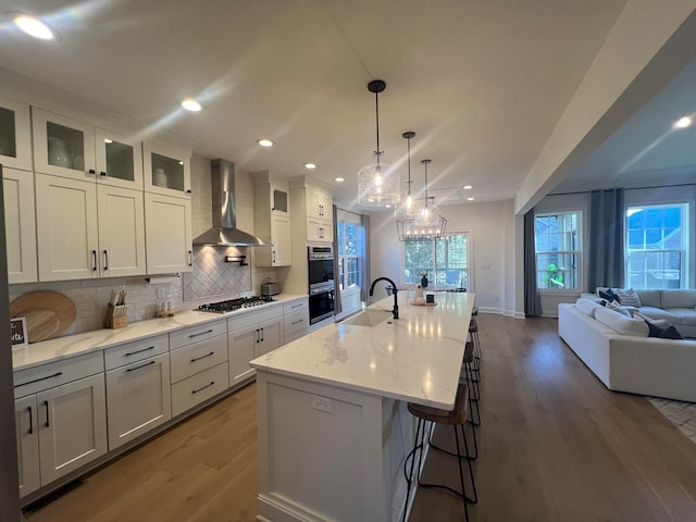 kitchen featuring wall chimney exhaust hood, stainless steel appliances, light stone counters, hardwood / wood-style floors, and a center island with sink