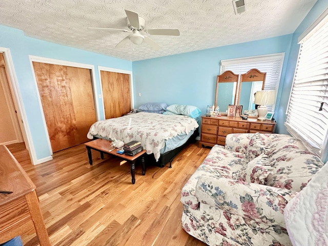 bedroom with multiple closets, light hardwood / wood-style flooring, a textured ceiling, and ceiling fan