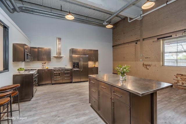 kitchen featuring stainless steel appliances, light hardwood / wood-style floors, a center island, a towering ceiling, and wall chimney range hood