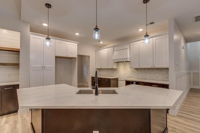 kitchen with custom exhaust hood, hanging light fixtures, a center island with sink, white cabinetry, and sink