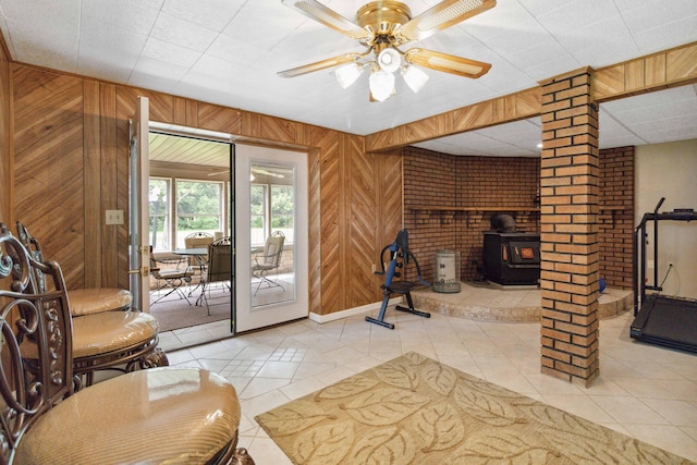 living room featuring ceiling fan, light tile patterned floors, wood walls, ornate columns, and a wood stove