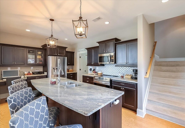 kitchen featuring sink, light wood-type flooring, an island with sink, appliances with stainless steel finishes, and decorative light fixtures