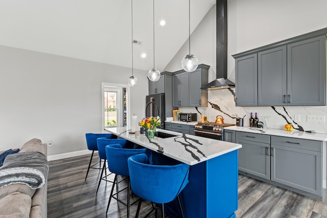 kitchen with gray cabinetry, wall chimney exhaust hood, an island with sink, pendant lighting, and appliances with stainless steel finishes