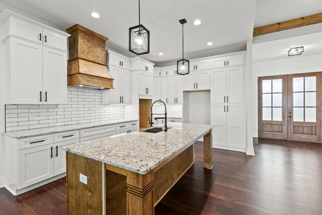 kitchen featuring custom exhaust hood, an island with sink, pendant lighting, and white cabinetry