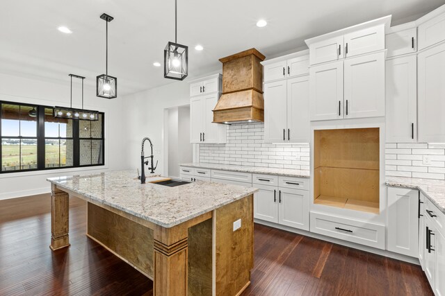 kitchen with light stone counters, hanging light fixtures, beamed ceiling, a kitchen island with sink, and white cabinets