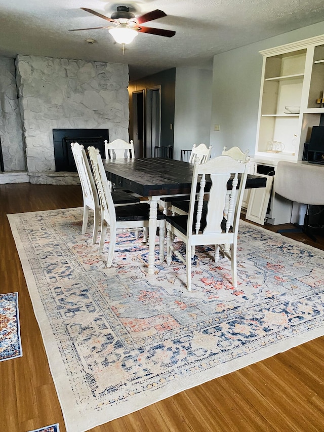 dining area featuring a stone fireplace, ceiling fan, wood-type flooring, and a textured ceiling