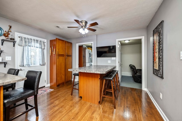 kitchen featuring ceiling fan, a kitchen breakfast bar, a textured ceiling, light hardwood / wood-style floors, and stacked washer and clothes dryer