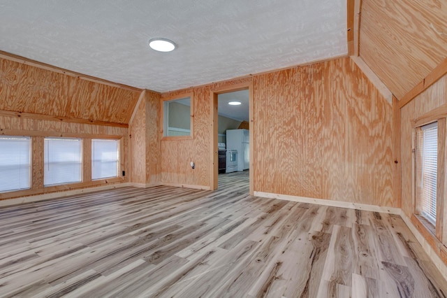 empty room featuring light hardwood / wood-style floors, wood walls, a textured ceiling, and vaulted ceiling