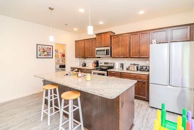 kitchen with light hardwood / wood-style floors, a center island with sink, appliances with stainless steel finishes, hanging light fixtures, and decorative backsplash