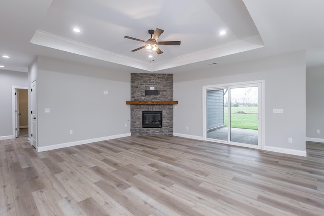 unfurnished living room featuring a tray ceiling, ceiling fan, crown molding, light hardwood / wood-style flooring, and a stone fireplace