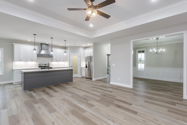 kitchen with stainless steel refrigerator with ice dispenser, wall chimney range hood, white cabinets, hanging light fixtures, and an island with sink