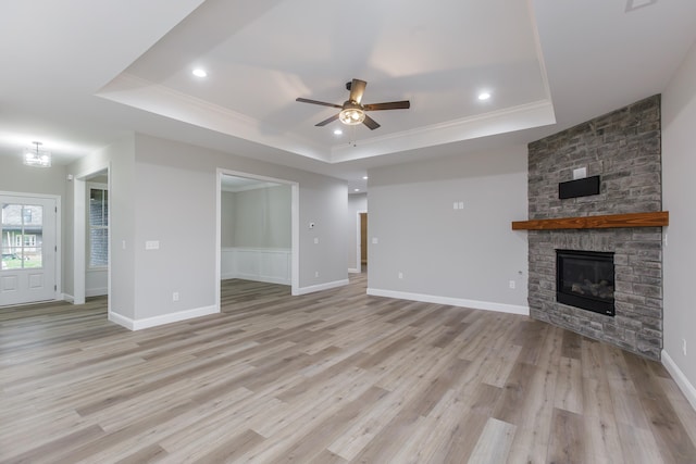 unfurnished living room with a stone fireplace, crown molding, ceiling fan, a tray ceiling, and light hardwood / wood-style floors