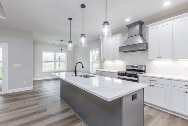 kitchen featuring sink, wall chimney range hood, stainless steel gas range, a center island with sink, and white cabinets