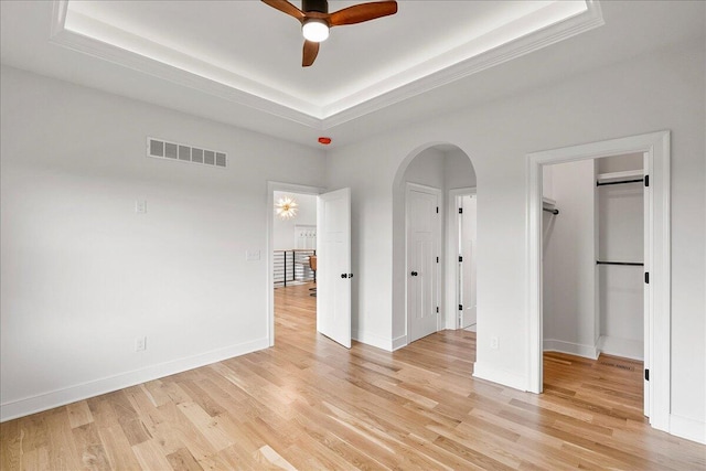 unfurnished bedroom with a tray ceiling, visible vents, light wood-style floors, arched walkways, and a spacious closet