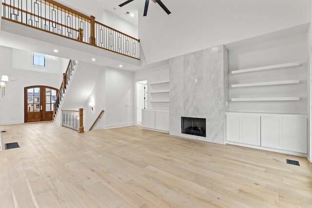 unfurnished living room with ceiling fan, a high ceiling, built in features, a high end fireplace, and light wood-type flooring