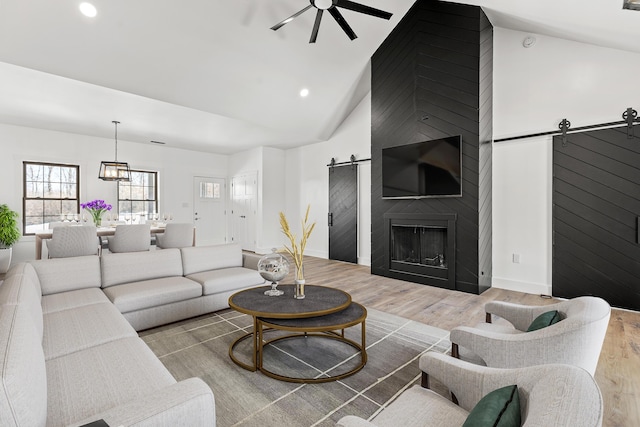 living room featuring ceiling fan, high vaulted ceiling, a fireplace, a barn door, and light wood-type flooring