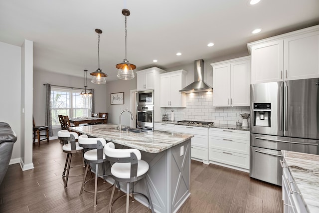 kitchen featuring wall chimney exhaust hood, light stone countertops, stainless steel appliances, and an island with sink