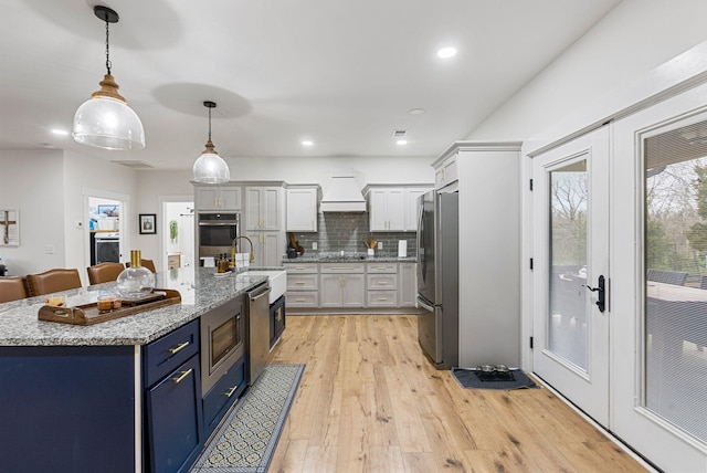 kitchen with light wood finished floors, tasteful backsplash, custom range hood, appliances with stainless steel finishes, and blue cabinets