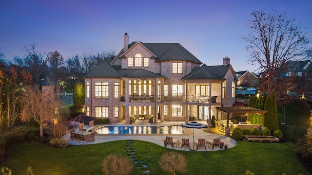 back house at dusk with a lawn, a patio area, a balcony, and a swimming pool with hot tub