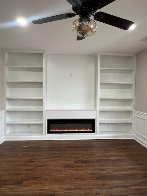 unfurnished living room with ceiling fan, a lit fireplace, built in shelves, and dark wood-style flooring