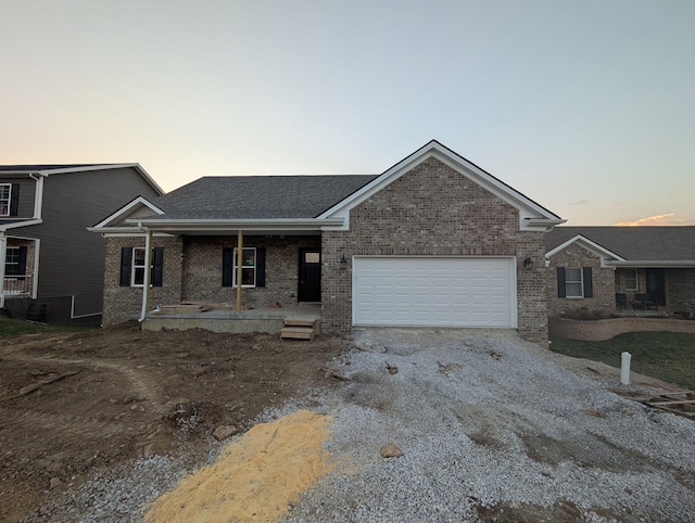 ranch-style home featuring brick siding, gravel driveway, roof with shingles, covered porch, and a garage