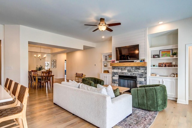 living room featuring built in shelves, a stone fireplace, light hardwood / wood-style flooring, and ceiling fan with notable chandelier