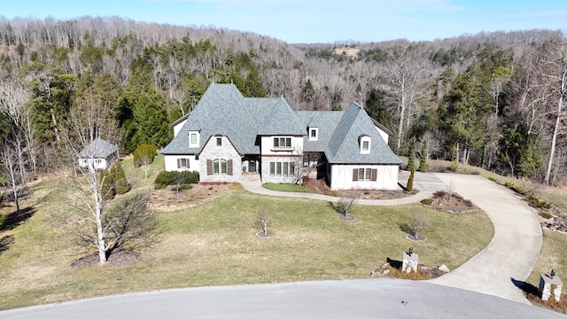 view of front of house featuring a front lawn, concrete driveway, stone siding, and a forest view