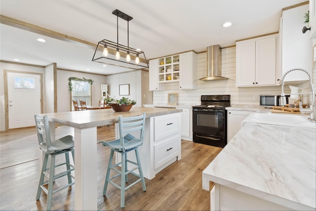 kitchen featuring black electric range, hanging light fixtures, a kitchen island, wall chimney range hood, and white cabinets