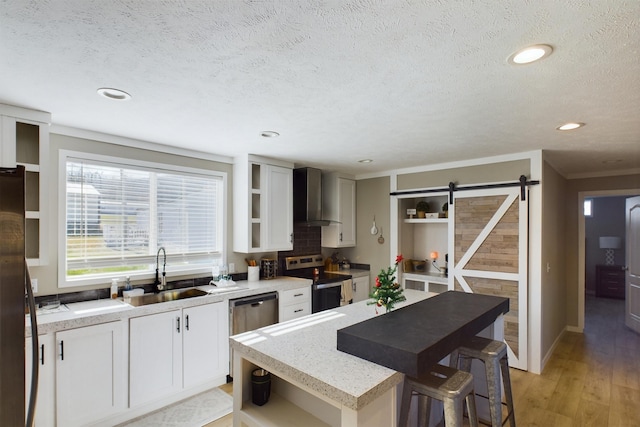 kitchen with wall chimney exhaust hood, stainless steel appliances, sink, a barn door, and white cabinets