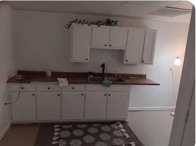 kitchen with dark countertops, baseboards, a drop ceiling, white cabinets, and a sink