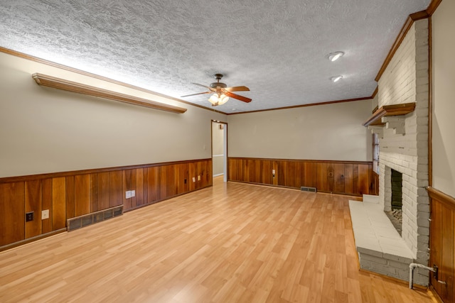 unfurnished living room with wood walls, a brick fireplace, ornamental molding, a textured ceiling, and light hardwood / wood-style floors