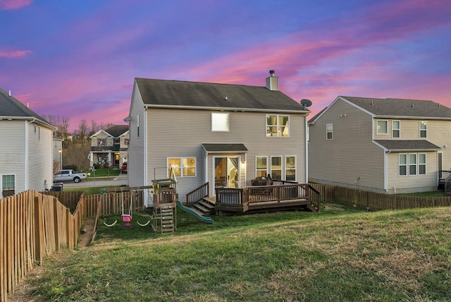 back house at dusk featuring a playground, a wooden deck, and a lawn