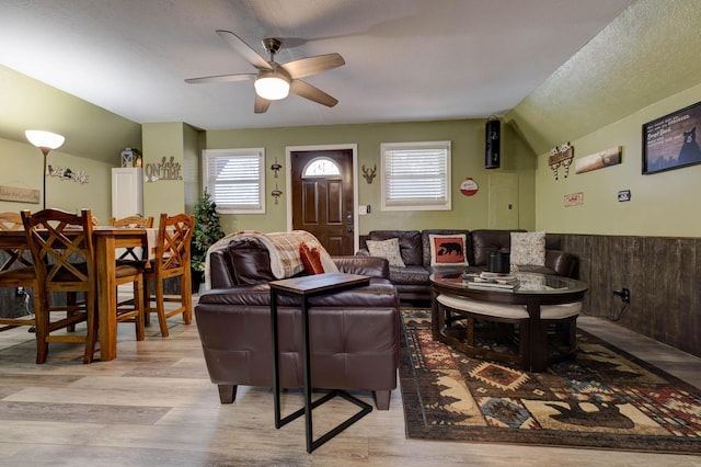 living room featuring ceiling fan, a textured ceiling, vaulted ceiling, wooden walls, and light wood-type flooring