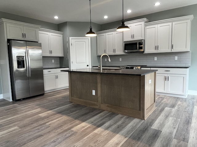 kitchen with light wood finished floors, stainless steel appliances, dark countertops, a sink, and an island with sink