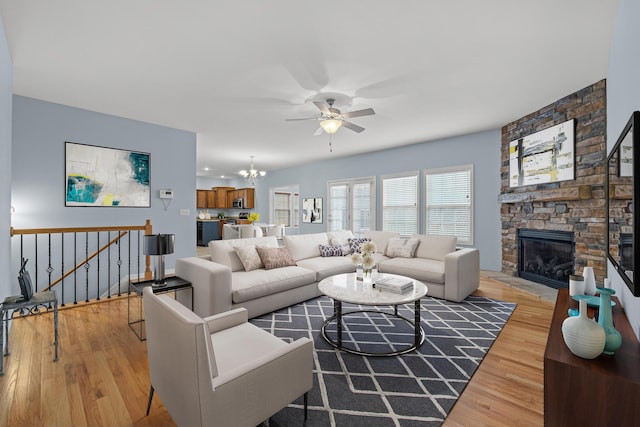 living room with light wood finished floors, a stone fireplace, and ceiling fan with notable chandelier