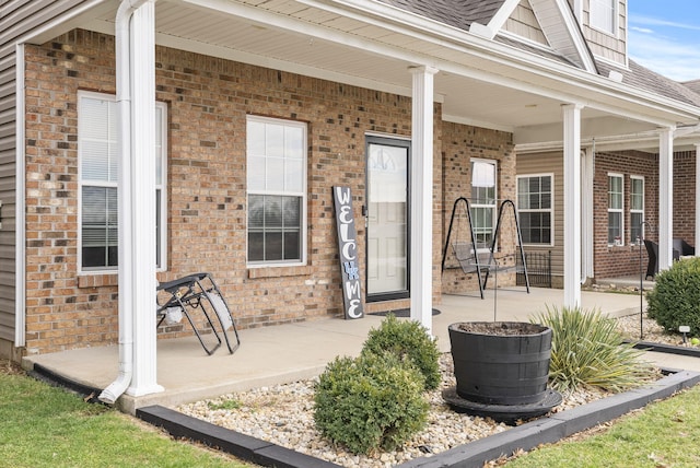 view of exterior entry with a porch, a shingled roof, and brick siding