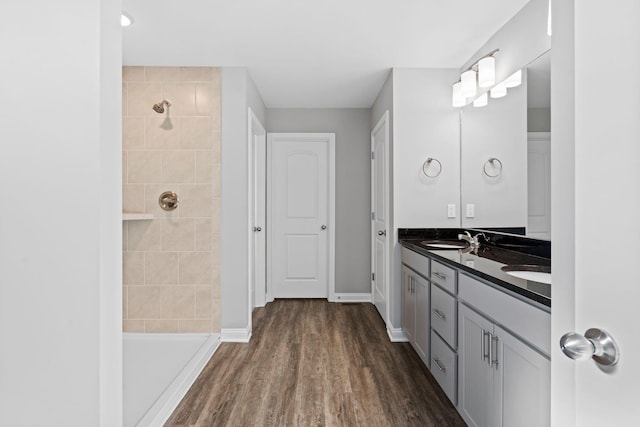full bathroom with a sink, a tile shower, double vanity, baseboards, and wood finished floors