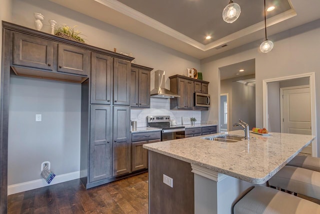 kitchen featuring sink, wall chimney range hood, a tray ceiling, a center island with sink, and appliances with stainless steel finishes