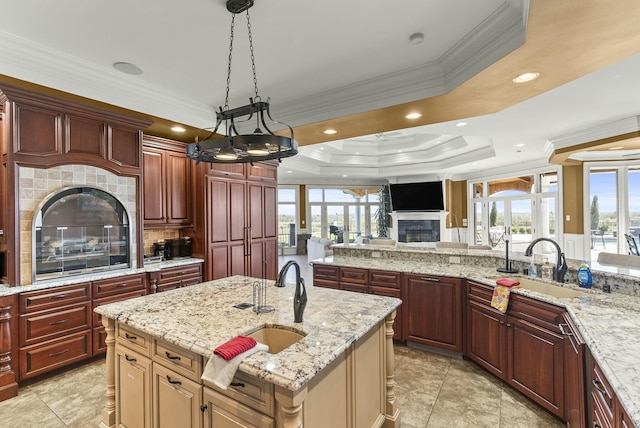 kitchen with pendant lighting, crown molding, light stone countertops, an island with sink, and a raised ceiling