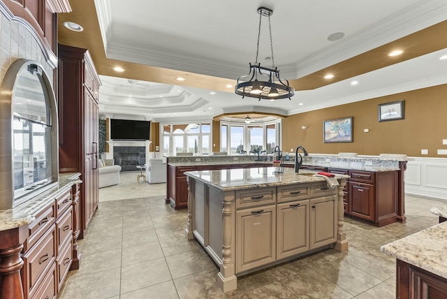 kitchen featuring a center island with sink, ornamental molding, a tray ceiling, pendant lighting, and light stone countertops