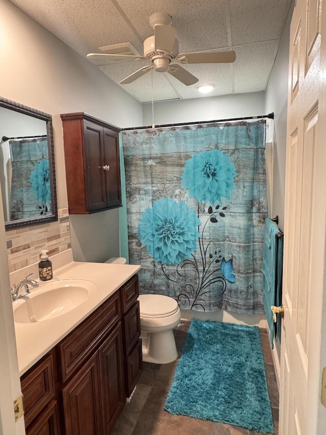 bathroom with ceiling fan, a drop ceiling, backsplash, toilet, and vanity