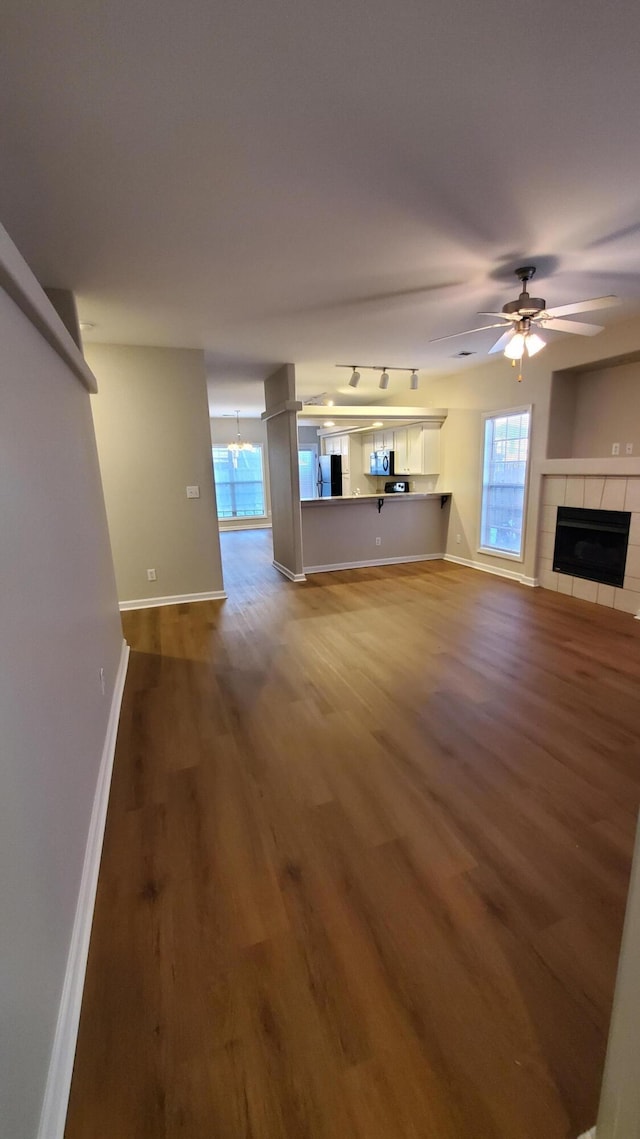 unfurnished living room featuring wood finished floors, baseboards, track lighting, a tiled fireplace, and ceiling fan with notable chandelier