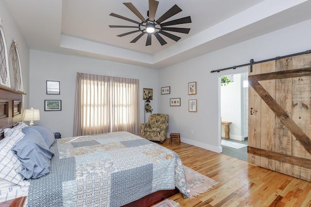 bedroom with ensuite bath, ceiling fan, a barn door, a tray ceiling, and hardwood / wood-style flooring