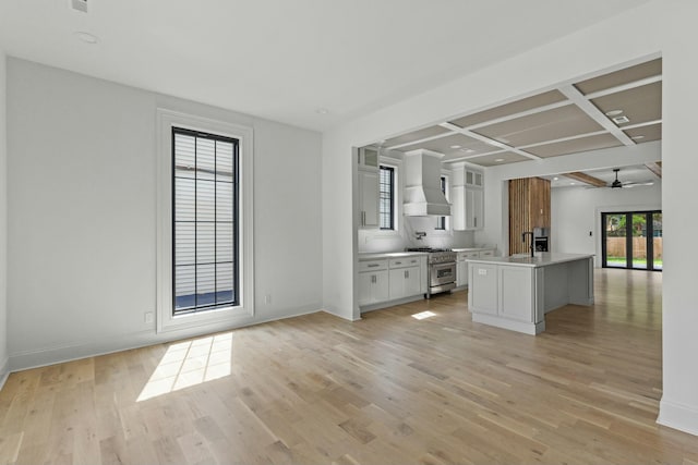 kitchen featuring coffered ceiling, a center island with sink, white cabinets, wall chimney exhaust hood, and high end stove